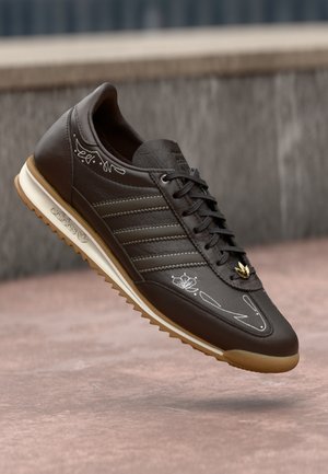 Donkerbruine Adidas sneaker met witte stiksels, gouden vetersieraad en lichtbruine zool, die boven een roze betonoppervlak zweeft.