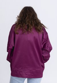 Oversized, paarse satijnen jacket met verlaagde schouders, elastische manchetten en een geometrisch gestikt zoomdetail. Achteraanzicht, gladde textuur.