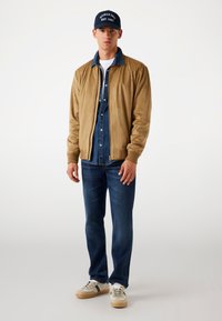 Bruin suède bomberjack over een denim shirt, donkere jeans en beige sneakers, aangevuld met een marineblauwe pet met witte tekst.