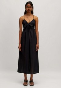 Schwarzes Maxikleid mit einer gebundenen Frontdetail und schmalen Trägern, gefertigt aus leichtem Stoff. Das Modell steht barfuß und betont den Schwung des Kleides.