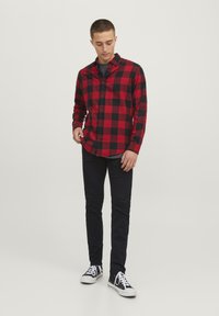Chemise en flanelle à carreaux rouges et noirs avec des boutons, manches longues et ourlet arrondi, portée avec un pantalon noir et des baskets noir et blanc.