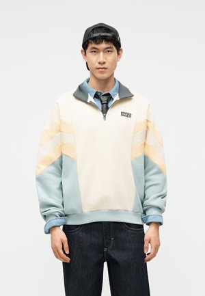 Fiatal férfi színes, negyedcipzáras pulóverben halványsárga és kék díszítéssel, farmeringben, fekete nyakkendővel, sötét farmerral és fekete sapkával, amelyet hátrafelé visel.