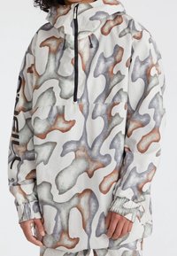 Leichter Pullover-Jacke mit weißem Grund; zeichnet sich durch ein abstraktes Tarnmuster in Grau, Rost und Grün aus. Enthält einen Reißverschluss vorne und eine Kapuze.