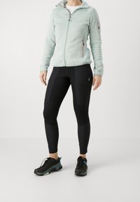 Femme portant une veste polaire zippée vert clair, des leggings noirs avec poches latérales, et des chaussures de randonnée noires, debout sur un fond uni.