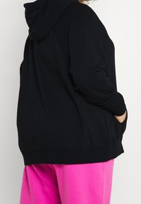 Person i svart hoodie med händerna i fickorna, kombinerad med ljusrosa byxor mot en enkel ljus bakgrund.