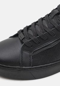 Baskets noires en cuir lisse avec des panneaux latéraux texturés, des lacets plats tissés, et une semelle en caoutchouc avec un motif géométrique subtil.