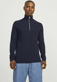 Navy-blau gerippter Rollkragenpullover mit einem Reißverschluss bis zur Viertellänge, schmaler Schnitt und langen Ärmeln, kombiniert mit hellblauen Jeans.