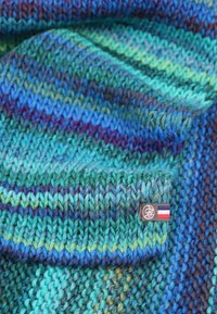 Écharpe tricotée avec des nuances variées de bleu et de vert, présentant un motif texturé et épais, ainsi qu'une étiquette en tissu avec un logo et un détail de drapeau.