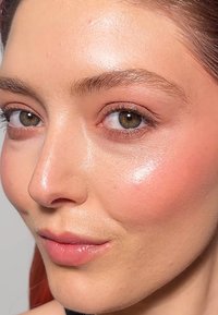 Primer plano de un rostro con piel suave y clara; ojos marrones; un sutil rubor rosa; labios brillantes; y cejas finas. El cabello es de un rojo oscuro.