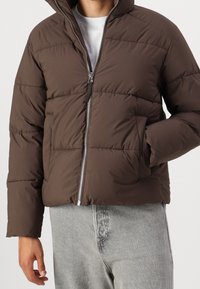 Hnědá prošívaná bunda s kapucí, stříbrným zipem, elastickými manžetami a diagonálním prošíváním. Nositelná přes šedé tričko a džíny.