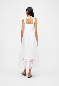 Γυναίκα που φοράει λευκό αμάνικο φόρεμα με φιόγκους στους ώμους και δαντέλα στο τελείωμα, όρθια ξυπόλητη μπροστά σε απλό φόντο.