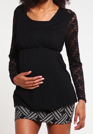 Gravid kvinde iført en sort top med blondeærmer og et mønstret sort-hvidt skørt, der holder sin mave med den ene hånd.