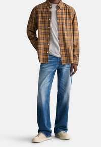 Brązowa i żółta koszula w kratę zapinana na guziki na szarym t-shircie, w połączeniu z jasnoniebieskimi dżinsami o prostym kroju i jasnobeżowymi sneakersami.