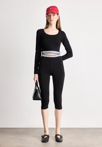 Žena nosi crnu majicu s dugim rukavima i Capri tajice s pojasom "Tommy Girl", crvenu kapu, crnu torbicu i crne balerinke.