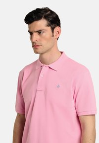 Polo de color rosa claro hecho de algodón, con un cuello clásico, mangas cortas y un pequeño logo azul en el pecho.