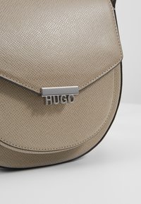Beżowa torebka typu crossbody z fakturą, z metalowym zapięciem w kolorze srebrnym z logo 'HUGO', o zaokrąglonym kształcie, z cienkim czarnym wykończeniem wzdłuż krawędzi.