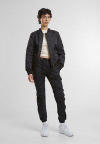 Schwarze Bomberjacke mit Reißverschluss, gerippten Bündchen und Saum. Kombiniert mit schwarzen Cargo-Hosen, weißen Sneakers und einem hellen Oberteil.