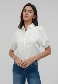 Fehér puff-ujjú blúz gombos elülső résszel, galléros nyakkal és rugalmas mandzsettákkal. Sötétkék farmernadrággal párosítva.