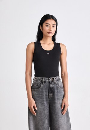 Γυναίκα με μακριά μαύρα μαλλιά, φοράει μαύρο αμάνικο τοπ και άνετο ψηλόμεσο γκρι τζιν, στέκεται μπροστά σε απλό λευκό φόντο.