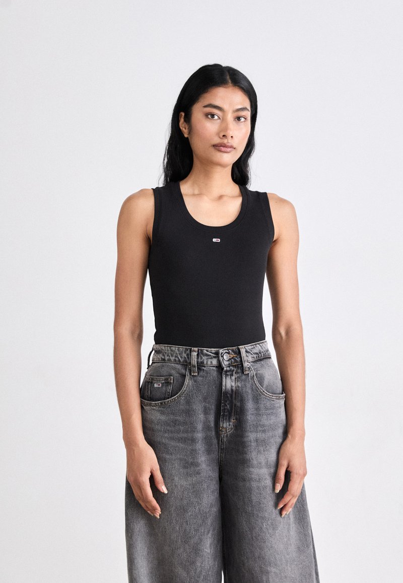 Kvinde med langt sort hår iført en sort ærmeløs top og højtaljede løse grå jeans, stående mod en ensfarvet hvid baggrund.