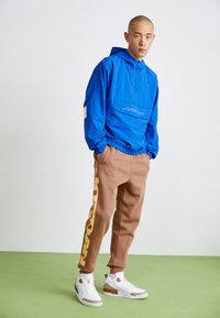 Férfi kék Jordan kapucnis pulóverben, barna melegítőnadrágban sárga felirattal és fehér tornacipőben áll egy zöld padlón, fehér fal előtt.