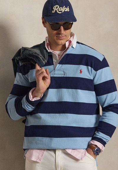 Homme portant un maillot de rugby rayé bleu marine et bleu clair, une chemise rose boutonnée en dessous, une casquette bleu marine avec le logo "Ralph", des lunettes de soleil, et portant une veste en jean.
