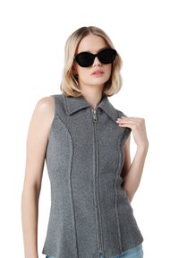 Vest tricotat gri fără mâneci, cu guler și fermoar frontal. Dispune de cusături texturate și o margine evazată. Modelul poartă ochelari de soare negri tip cat-eye.