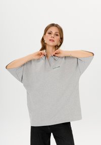 Helle graue Oversized-Polohemd aus Baumwollmischung, mit Kragen, kurzen Ärmeln und einem grünen Logo auf der Vorderseite. Glatte Textur.