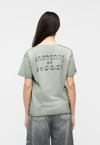 Άτομο με μακριά σκούρα μαλλιά που φοράει ξεθωριασμένο πράσινο μπλουζάκι με το κείμενο «PROPERTY OF DIESEL» και ξεθωριασμένο γκρι τζιν, φαίνεται από πίσω.