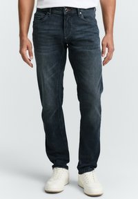 Vaqueros de mezclilla azul oscuro con corte recto ajustado. Presenta cinco bolsillos, un ligero desvanecimiento y un detalle de logotipo en la parte frontal. Combinados con zapatillas blancas.