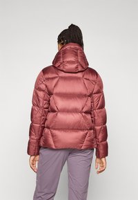 Una persona con trenzas lleva una chaqueta acolchada burdeos con capucha, de espaldas contra un fondo claro y liso.