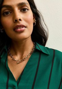 Gouden ketting met een verlengd schakeldesign, voorzien van een sluiting met een haak, gedragen over een groen gestreept blouse.