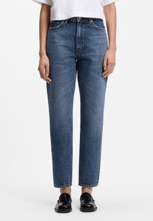 Person iført højtsiddende, lige ben blå jeans parret med sorte loafers og en hvid cropped top, stående mod en ensfarvet baggrund.