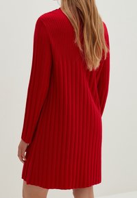 Robe-pull rouge côtelée avec manches longues, présentant une texture verticale. La coupe légèrement évasée arrive à mi-cuisse.