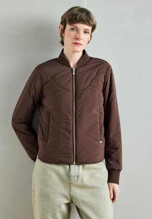 Καφέ μπουφάν bomber με ραφές, ριμπ γιακά, μπροστινές τσέπες και ασημένια λεπτομέρεια φερμουάρ, συνδυασμένο με ανοιχτόχρωμο τζιν παντελόνι.