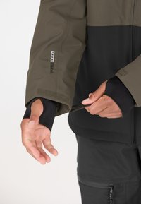 Wasserabweisende grün-schwarze Jacke mit Klettverschlüssen an den Ärmeln, die eine matte Textur aufweist und elastische Daumenschlaufen für einen verbesserten Sitz und Komfort bietet.