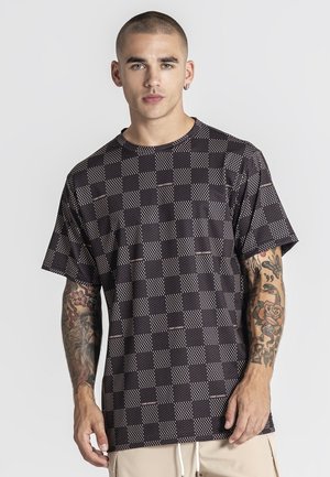 Giovane uomo con capelli corti e tatuaggi indossa una camicia a maniche corte a quadri nera e grigia e pantaloni beige su uno sfondo bianco.