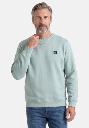Midaldrende mand med gråt hår og skæg iført lyseblå sweatshirt og blå jeans, der rører ved kraven med én hånd.