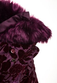 Collar de pelo sintético morado con ribete de satén sobre un abrigo de terciopelo morado que presenta patrones florales y botones decorativos. Textura y color ricos.