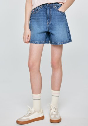 Osoba ubrana w niebieskie szorty z denimu, białe skarpetki z szarymi i różowymi paskami oraz białe tenisówki na jasnym, jednolitym tle.