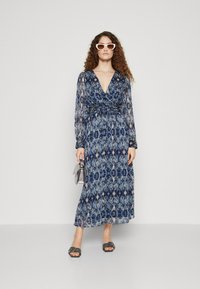 Rochie lungă, cu un model albastru, decolteu adânc în formă de V, mâneci lungi și talie elastică. Modelul ține o poșetă argintie și poartă ochelari de soare albi.