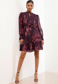 Donna che indossa un vestito floreale viola scuro con maniche lunghe trasparenti e orlo a volant, in piedi con sicurezza contro un muro bianco.