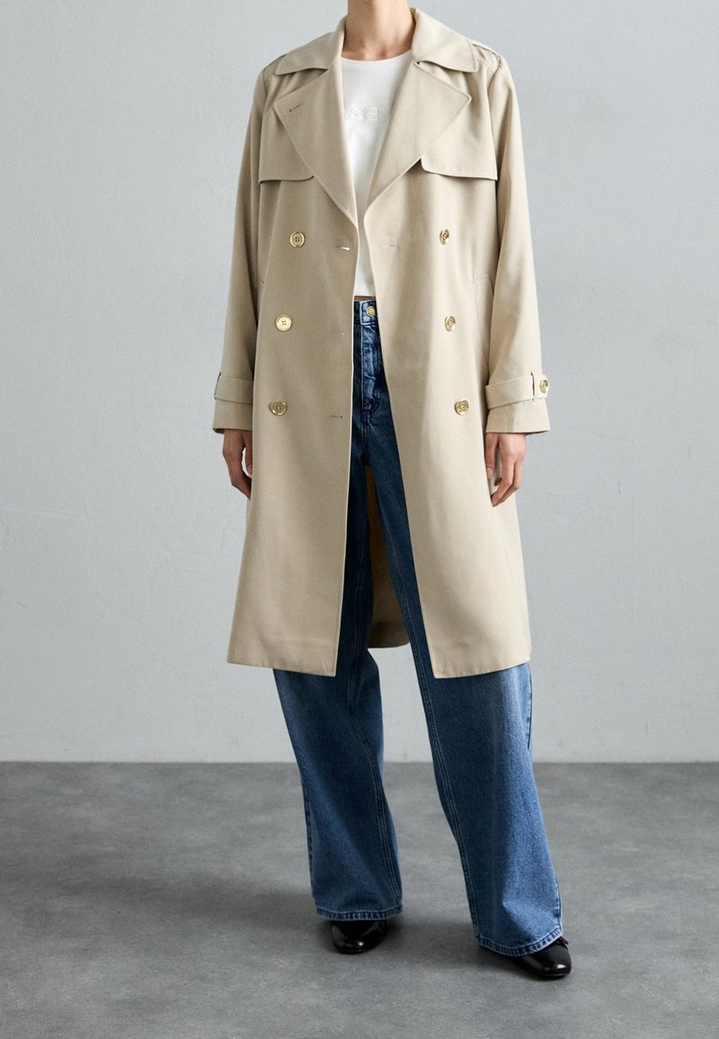 Personne portant un trench-coat beige à double boutonnage sur un haut blanc, un jean bleu à jambes larges et des chaussures noires, debout sur un sol gris.