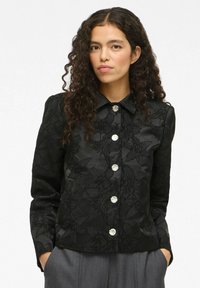 Chemise en satin noir avec texture florale, manches longues et col pointu. Présente de grands boutons transparents et une coupe ajustée.