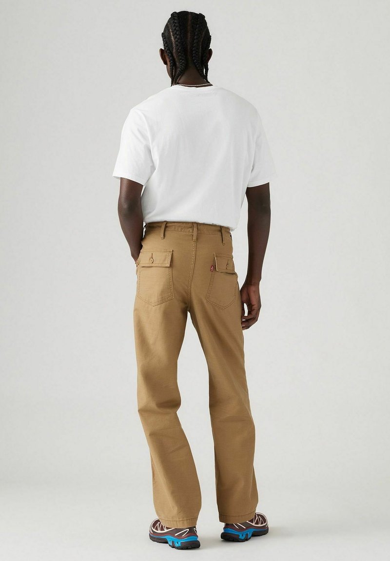 Pantalones cargo marrones con bolsillos visibles, combinados con una camiseta blanca lisa. Hombre de pie con cabello trenzado, mostrando un diseño de corte relajado.