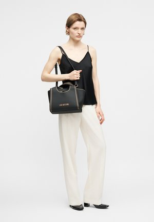 Γυναίκα που φοράει μαύρο αμάνικο τοπ και λευκό παντελόνι, κρατώντας μια μαύρη τσάντα Love Moschino με χρυστά γαζιά στον ώμο της.