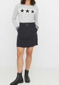 Grauer Pullover mit langen Ärmeln und schwarzen Sterne-Applikationen, kombiniert mit einem schwarzen Rock mit Taschen und einem Gürtel, abgerundet mit schwarzen Knöchelstiefeln.