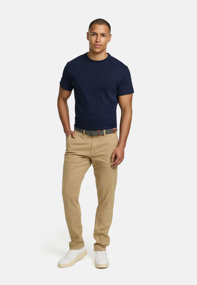 Navy cotton t-shirt paired with beige pants and white sneakers. The model wears a patterned belt and stands with hands in pockets.
