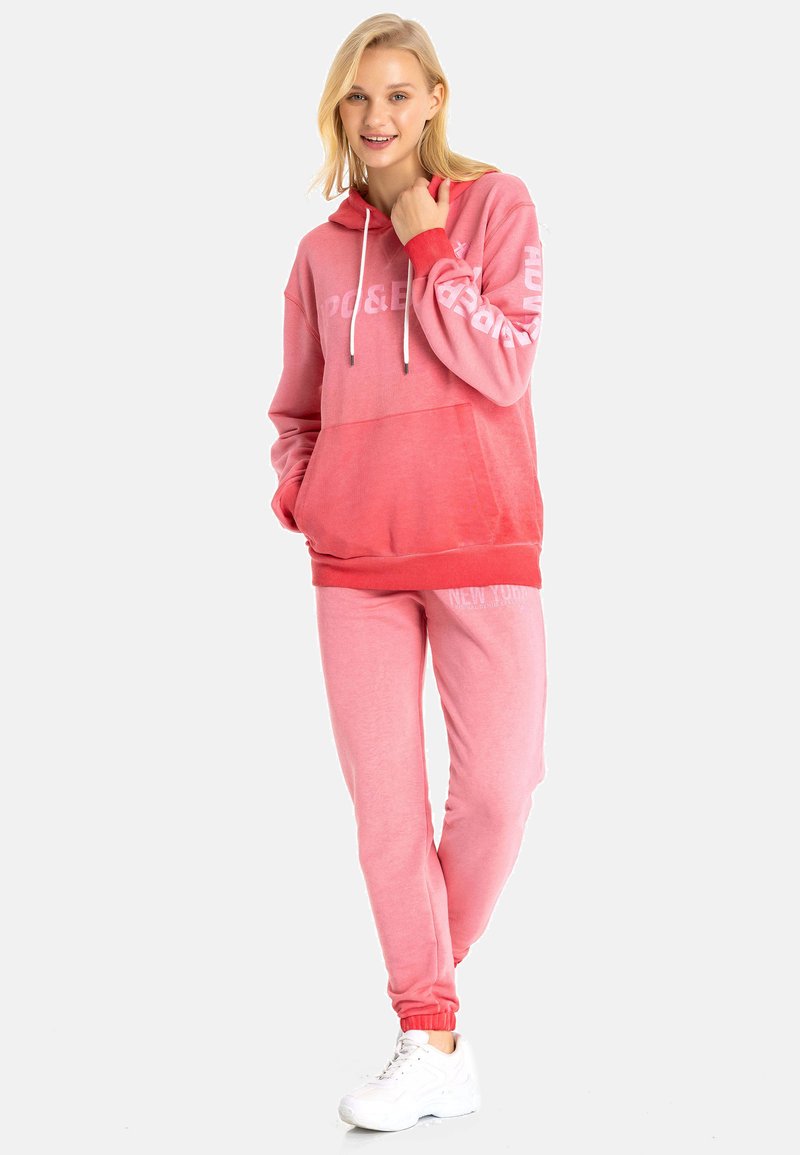 Rosa Kapuzenpullover und passende Jogginghose aus weichem Stoff, mit weißen Text und einer Kordelzugkapuze. Betonte Bündchen und entspannter Schnitt.