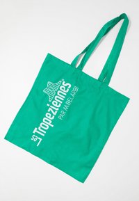 Bolsa de algodón verde con dos asas. Presenta texto blanco y un logotipo en un lado. Diseño simple, forma rectangular y material ligero.